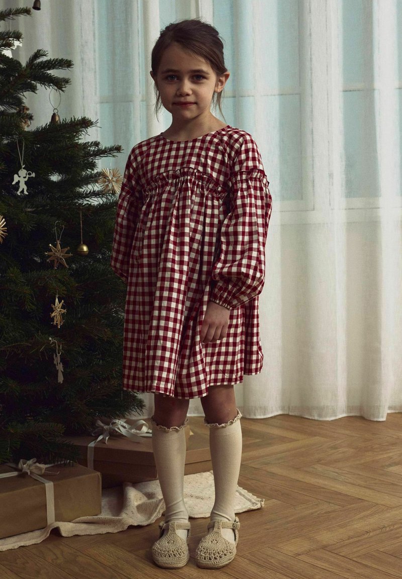 Vestido a cuadros rojos y blancos con mangas abullonadas y cintura fruncida, combinado con medias crema hasta la rodilla y zapatos de mimbre beige.