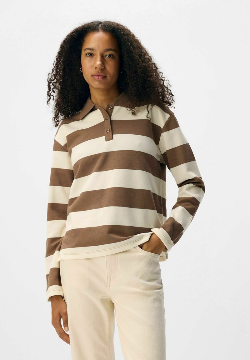 Polo de manga larga a rayas en color blanco roto y marrón. Presenta un cuello abotonado y una textura suave. Combinado con pantalones de color claro.