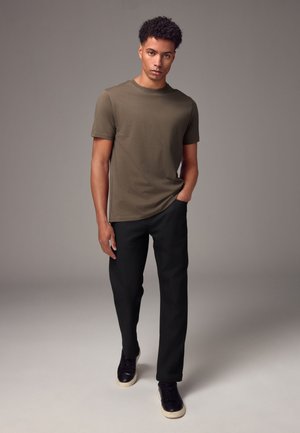 Giovane uomo con capelli ricci che indossa una t-shirt verde oliva, pantaloni neri e sneakers nere, in piedi con una mano in tasca.