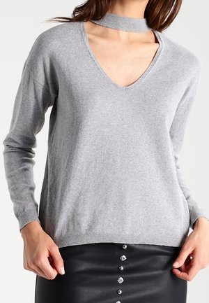 Mujer con un suéter de pico gris claro y un collar choker a juego, y una falda de cuero negra con botones plateados.