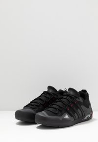 Chaussures de sport noires avec un dessus en maille texturée, capuchon en caoutchouc à l'avant, et accents rouges. Elles possèdent un design à lacets et une semelle rayée pour une meilleure traction.