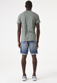 T-shirt verde con motivo a foglie, abbinato a pantaloni shorts in denim blu con risvolti e due tasche sul retro. Sneakers con accenti multicolori.