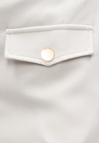 Tissu gris clair avec une poche à rabat ornée d'un bouton rond en or. Le matériau semble lisse avec une texture subtile.