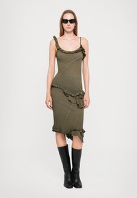 Rochie verde măslinie cu accente volă, silueta ajustată și tiv asimetric. Modelul poartă cizme negre până la genunchi și ochelari de soare.