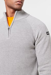 Szary sweter dzianinowy z kołnierzem na pół suwaka, teksturowaną tkaniną, prążkowanym wzorem na ramionach oraz naszywką z logo na lewym ramieniu. Wewnętrzna warstwa jest pomarańczowa.