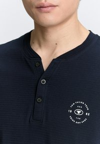Tamsiai mėlynas medvilninis trumpomis rankovėmis henley marškinėliai su trimis juodais mygtukais, banguota apykakle ir baltu apskritu logotipu ant krūtinės.