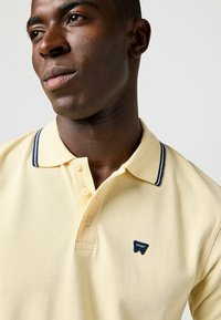 Polohemd aus hellgelbem Stoff mit strukturierter Oberfläche, kragen in marineblau mit Streifen, drei Knöpfen und einem blauen dreieckigen Wrangler-Logo.