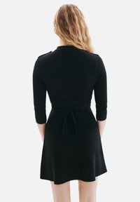 Robe noire avec des manches trois-quarts, taille cintrée et jupe évasée. Présente une ceinture à nouer à l'arrière et une texture en tricot lisse.