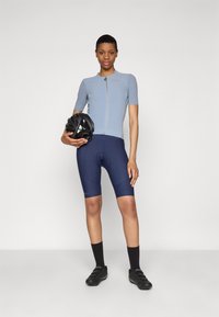 Radsport-Outfit bestehend aus einem hellblauen, kurzärmeligen, mit Reißverschluss versehenen Trikot und navyblauen Radsportshorts, mit einem Helm in der Hand. Schwarze Radschuhe.