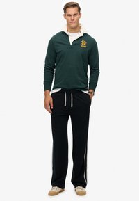 Man in een donkergroen rugbyshirt met witte kraag, zwarte broek met trekkoord en zijstrepen, beige sneakers en een polshorloge, die recht vooruit staat.