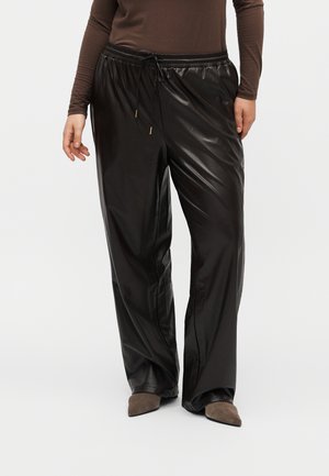 Personne portant un pantalon large noir brillant avec une taille élastique et un cordon, associé à des chaussures pointues marron.