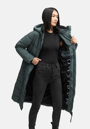 Chaqueta de plumas larga en verde oscuro con capucha. Presenta un diseño acolchado, cierre de cremallera y forro negro con un logo de la marca. Llevada abierta sobre ropa negra.
