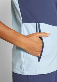 Hand inserting into zipped side pocket of light blue and navy short-sleeve jacket with matching navy pants visible below.