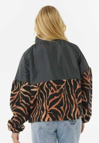 Chaqueta de forro polar a rayas negra y naranja con un panel superior negro brillante, que presenta mangas largas y puños elásticos. La vista trasera muestra un contraste en el diseño.