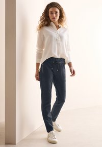 Biała koszula z długim rękawem i złożonym kołnierzykiem, połączona z granatowymi spodniami dresowymi z kieszeniami i sznurkiem w talii. Całość dopełniają białe sneakersy.