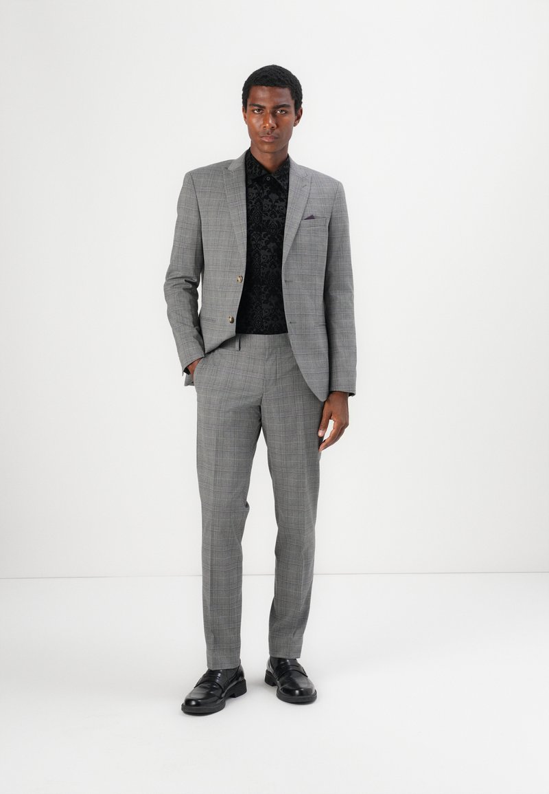 Grijs geruit pak bestaande uit een getailleerde jas en bijpassende broek, gedragen over een gestructureerd zwart overhemd, aangevuld met zwarte schoenen.