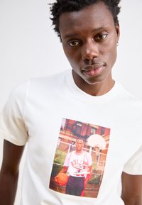 Λευκό μπλουζάκι με γραφική εκτύπωση μπροστά ενός άνδρα καλαθοσφαιριστή να κρατάει μια μπάλα, με φόντο γήπεδο μπάσκετ και αστικά στοιχεία.