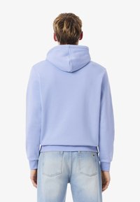 Sudadera azul claro con un bolsillo tipo canguro y puños acanalados. La tela parece suave; presenta una capucha amplia y un diseño minimalista. Se lleva con unos pantalones vaqueros claros.