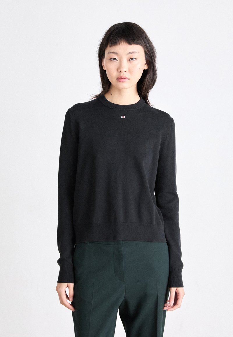 Pull noir à col rond en tissu tricoté, avec un petit logo sur la poitrine, des manches longues et un ourlet droit. Porté avec un pantalon vert.