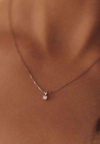 Collier en chaîne argentée avec un petit pendentif en pierre précieuse carrée et transparente. La chaîne est fine avec des maillons, sur un fond de teint neutre.