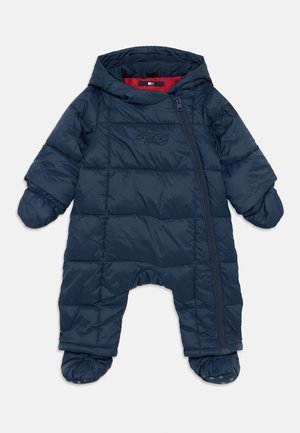 Combinaison de neige rembourrée de couleur bleu marine avec fermeture éclair, capuche détachable et pieds recouverts. Présente un motif matelassé et un logo sur la poitrine.