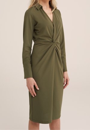 Olijfgroene jurk met een gedraaide voorkant, lange mouwen en kraag. Gemaakt van een gladde stof, knielang met een getailleerd silhouet.