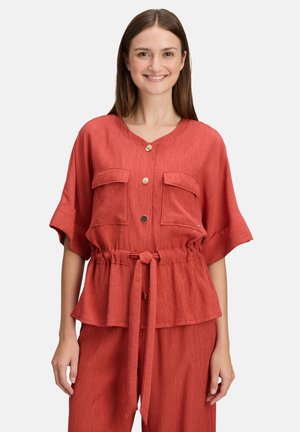 Rode blouse met textuur, korte mouwen, ronde halslijn, gouden knopen en twee borstzakken. Voorzien van een verstelbare taille met trekkoord.