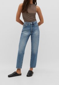 Mujer vestida con una blusa sin mangas color topo, jeans de cintura alta en un tono azul claro y zuecos negros sin cordones con tachuelas doradas, de pie contra un fondo liso.