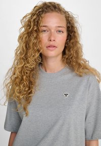 Camiseta de algodón gris con mangas cortas, cuello redondo y un pequeño emblema de abeja en blanco y negro en el lado izquierdo del pecho.