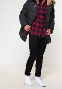 Czarna kurtka puchowa z kołnierzem z futerka syntetycznego, czerwona i granatowa koszula w kratę, czarne spodnie oraz białe trampki. Skupienie na nakładaniu warstw i kontraście kolorów.