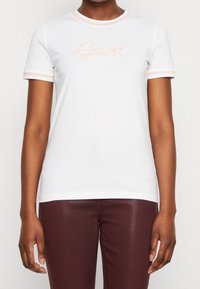 Vit kortärmad T-shirt med ljusrosa detaljer och "Guess"-logotyp, bärs med bordeauxfärgade tajta byxor, visar torso och armar.