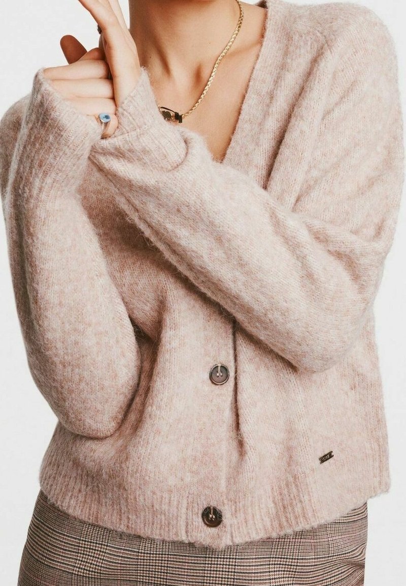 Personne portant un cardigan rose pâle boutonné et un pantalon à carreaux, montrant une bague avec une pierre bleue à son doigt et un collier chaîne en or.