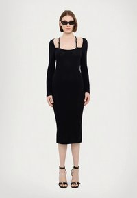Czarna dzianinowa sukienka midi z długimi rękawami, kwadratowym dekoltem i ozdobnymi paskami. Połączona z czarnymi sandałami na paski i pasującymi okularami przeciwsłonecznymi.