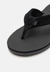 Tongs noires en caoutchouc, avec une semelle texturée et des fines sangles. Les sangles présentent des coutures et une petite étiquette de logo.