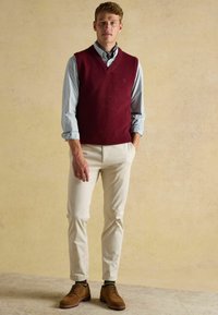 Burgundowy wełniany kamizelka nałożona na jasnoniebieską, w paski koszulę, w połączeniu z kremowymi spodniami i brązowymi zamszowymi butami, prezentująca klasyczny, casualowy wygląd.