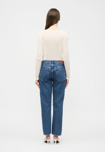 Woman with long brown hair wearing a sheer cream long-sleeve top, high-waisted blue jeans, and white heeled shoes, standing facing away.