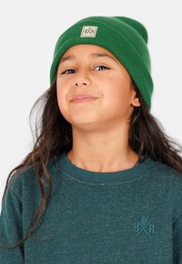Grön stickad mössa med vikta kanter och en fyrkantig lapp med en logotyp. Modellen har på sig en mörkgrön strukturerad sweatshirt under.