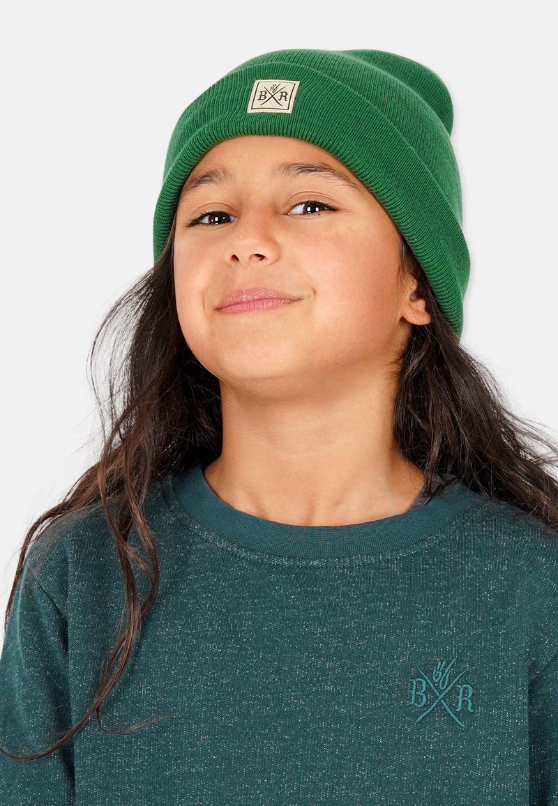 Grön stickad mössa med vikta kanter och en fyrkantig lapp med en logotyp. Modellen har på sig en mörkgrön strukturerad sweatshirt under.
