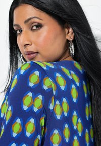 Blau gemusterte Bluse mit kreisförmigen Designs in Grün, Hellblau und Braun. Glatte Stoffqualität mit lockerer Passform und kurzen Ärmeln.