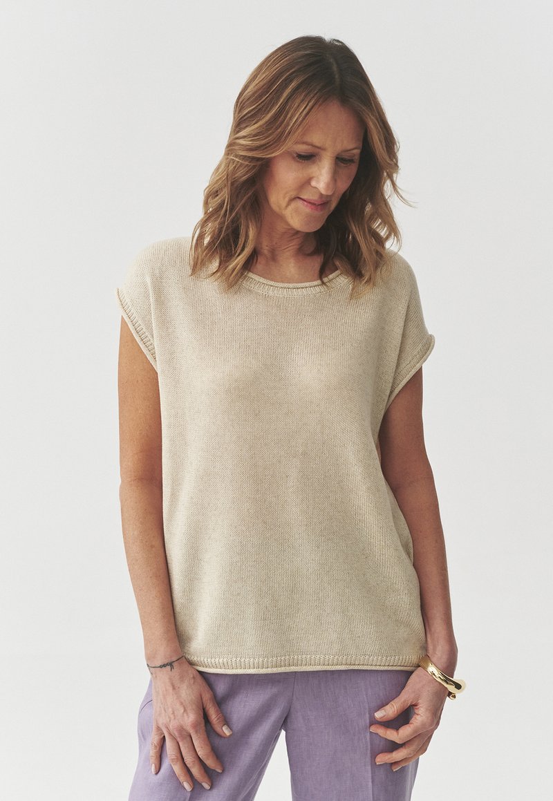 Beige pletený top s krátkým rukávem, kulatým výstřihem a texturovanou tkaninou. Volný střih, nošený s světle fialovými kalhotami s minimalistickým designem.