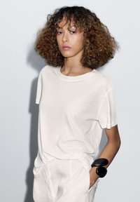 Hvid bomulds t-shirt med korte ærmer, der har en afslappet pasform. Et sort armbånd fremhæver håndleddet. Enkel og minimalistisk design.