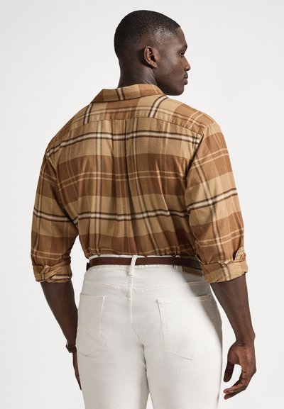 Rjava karo s srajco z gumbi in zavihanimi rokavi, izdelana iz tkanine z gladko teksturo. Kombinirana z belimi hlačami in rjavim pasom.