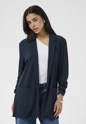 Młoda kobieta z długimi ciemnymi włosami, ubrana w granatową marynarkę zapinaną na otwarto, biały prążkowany top, niebieskie dżinsy, perłowy kolczyk i srebrną bransoletkę, pozująca.