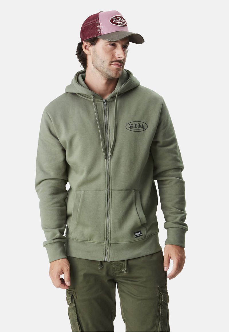 Sweatshirt zippé vert olive avec poches avant et patch logo. Associé à un pantalon cargo olive. Le chapeau a une partie arrière en maille bordeaux et une partie avant beige.
