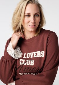 Bruine trui met witte tekst "LOVERS CLUB." Voorzien van kanten accenten op de mouwen en een ontspannen ontwerp. Effen achtergrond.