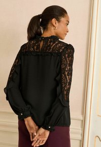 Fekete blúz csipkés részletekkel a vállakon, fodros ujjakkal és gombos mandzsettákkal. Kulcslyuk alakú nyílás a hátán.