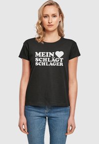 Sort bomulds T-shirt med rund halsudskæring, prydet med hvid tekst: "MEIN ♥ SCHLÄGT SCHLAGER." Korte ærmer og en afslappet pasform.