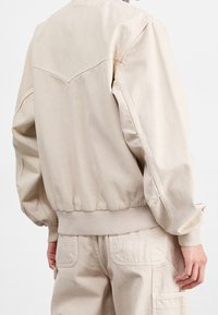 Veste bomber beige en mélange de coton avec ourlet et poignets côtelés, dotée d'un design yoke au dos et d'une taille élastiquée pour un ajustement parfait.