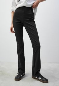 Pantalon noir évasé en tissu extensible, avec une taille haute et une texture lisse. Associé à des baskets noires.