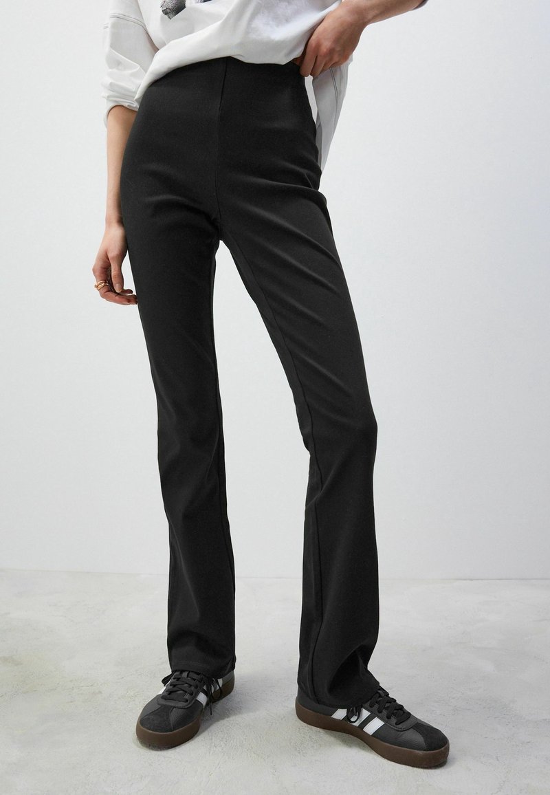 Pantalon noir évasé en tissu extensible, avec une taille haute et une texture lisse. Associé à des baskets noires.